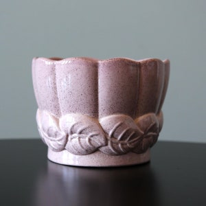 Könnte beinhalten: Ein rosafarbener Keramiktopf mit einem gewellten Rand und einem Blattdesign um die Basis.