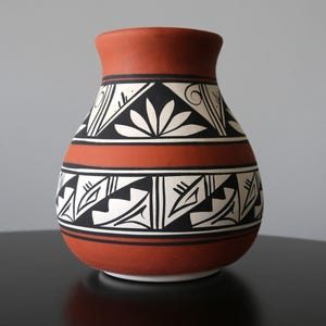 Może przedstawiać: Terakotowy wazon o szerokiej podstawie i wąskiej szyi. Wazon zdobiony czarno-białymi geometrycznymi i kwiatowymi wzorami. Wzory ułożone są w pasy wokół korpusu, tworząc efekt dekoracyjny. Wysokość około 20 cm.