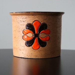 Peut inclure: Pot cylindrique en céramique avec un extérieur texturé de couleur beige. Le pot présente un motif floral décoratif orange et noir. Le bord du pot est brun foncé. Le pot est posé sur une surface noire.