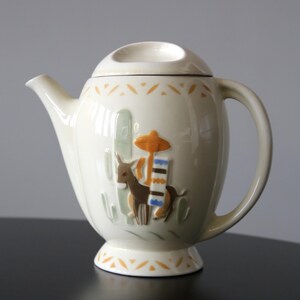 May include: Cream ceramic teapot with lid. The teapot has a raised relief design of a donkey, cactus, and sombrero. The handle and spout are curved. The base and rim have a repeating orange triangle pattern.