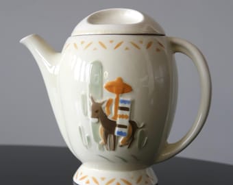 May include: Cream-colored ceramic teapot with a lid. The teapot features a raised relief design of a donkey, cactus, and a sombrero. The handle and spout are curved. The base and rim have a repeating orange triangle pattern.