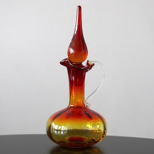 Puede incluir: Una jarra decorativa de vidrio con un tapón largo y puntiagudo. La jarra presenta un degradado de colores, que va desde una base amarillo dorado hasta un naranja vibrante y un rojo intenso en la parte superior. Tiene un asa de vidrio transparente.