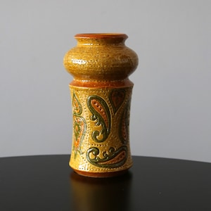 Op de afbeelding: Een bruine keramische vaas met een paisley-patroon in tinten groen, oranje en bruin. De vaas heeft een afgeronde bovenkant en een cilindrisch lichaam.