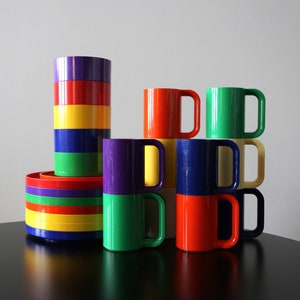 Może przedstawiać: Zestaw kolorowych plastikowych kubków i talerzy. Kubki są ułożone w kształcie piramidy, z górnym kubkiem w kolorze zielonym i żółtym. Talerze są ułożone w wieżę, z górnym talerzem w kolorze fioletowym i żółtym.