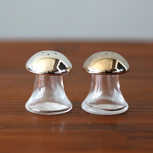 May include: A pair of mushroom-shaped salt and pepper shakers. The shakers have clear glass bodies and metallic silver-colored tops with small holes. They sit on a dark brown wooden surface.