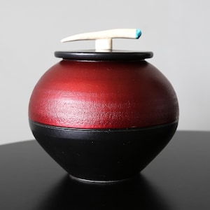 May include: A red and black ceramic jar with a white bone handle and a turquoise accent. The jar has a rounded shape and a smooth finish.