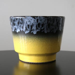 May include: A ceramic planter with a gradient design. The top rim is dark blue with a white, textured pattern, transitioning to black, then a bright yellow base. The planter has a stepped base and a smooth, glossy finish.