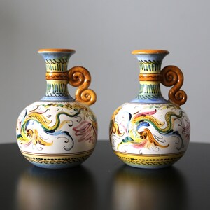 May include: Two ceramic vases with a blue, yellow, and white floral design. The vases have a handle that curls around the top.