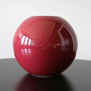 May include: A spherical vase with a glossy, deep red finish. The surface is smooth and reflective, showing light highlights across its rounded form. The vase features a circular opening at the top and a solid, uniform colour throughout its exterior.