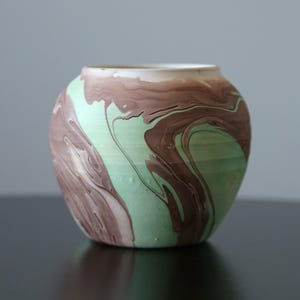 Puede incluir: Un jarrón redondo de cerámica con diseño veteado. El jarrón presenta una mezcla de tonos verde menta y marrón, creando un patrón arremolinado único. El borde del jarrón es de color crema claro. El jarrón está sobre una superficie oscura.