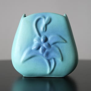 May include: A small, turquoise ceramic vase with a floral design. The vase has a rounded shape with a slightly flared top and a raised flower and leaf pattern in a darker shade of blue. The vase is smooth and glossy.