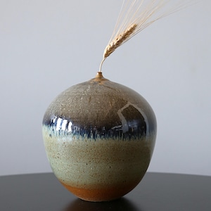 Könnte beinhalten: Eine handgefertigte Keramikvase mit runder Form und schmaler Öffnung. Die Vase weist einen Farbverlauf auf, der von Braun über Blau und Grün bis hin zu einem Hauch von Orange am Boden reicht. Ein einzelner Weizenhalm ist in die Vase gesteckt.