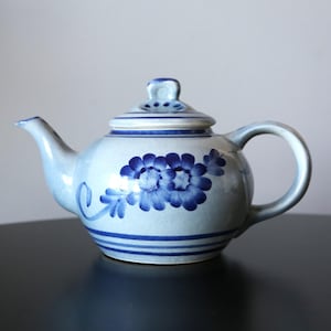 以下が含まれることがあります： 青と白のセラミック製のティーポットで、花柄が描かれています。ティーポットは丸いボディで、蓋の上にはノブが付いています。花柄は青色で、葉が付いたスタイリッシュな花が描かれています。