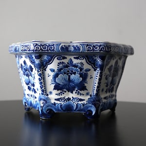 Puede incluir: Una jardinera de cerámica azul y blanca con un diseño floral. La jardinera tiene forma hexagonal con un borde festoneado y pequeños pies. El patrón floral azul se establece sobre un fondo blanco.