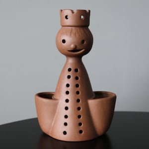 Op de afbeelding: Een bruin keramiek beeldje van een persoon met een kroon. Het beeldje heeft een glimlachend gezicht en een lichaam met gaten. Het beeldje staat op een bruine basis.