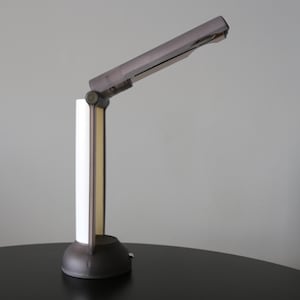 May include: A modern desk lamp with a dark gray finish. The lamp features a rectangular head and a vertical stem with a white panel. The lamp is positioned on a black surface.
