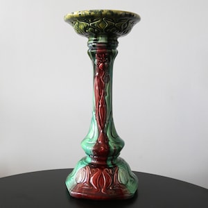 Könnte beinhalten: Ein dekorativer Keramiksockel in Grün, Rot und Braun. Der Sockel hat eine breite, gewellte Oberseite, einen schlanken, verzierten Stiel und einen ausgestellten Sockel. Das Design umfasst Blumen- und Blattmotive. Höhe ca. 30 cm.