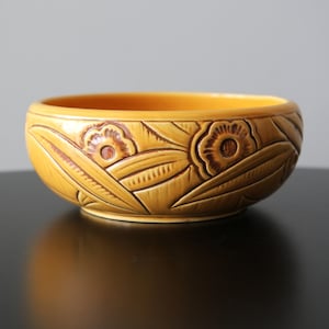 Puede incluir: Un cuenco de cerámica amarillo mostaza con un diseño floral y de hojas tallado en el exterior. El cuenco tiene forma redondeada y un acabado liso y brillante. Los detalles tallados son de un tono marrón más oscuro.