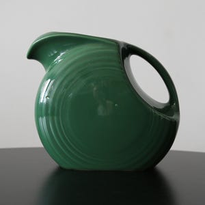 May include: A green ceramic pitcher with a circular body and a curved spout. The pitcher has a large handle and a glossy finish. The design features concentric rings around the body. The pitcher is sitting on a black surface.