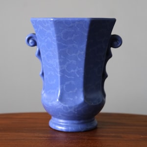 May include: A blue ceramic vase with a textured surface and a geometric design. The vase has two curved handles and a pedestal base. The vase is approximately 20 cm tall and 15 cm wide.