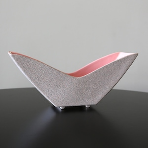 May include: A boat-shaped ceramic bowl with a textured, speckled grey exterior and a smooth, glossy pastel pink interior. The vessel features a pointed, angular silhouette with a wide opening and rests on small, subtle feet at the base.