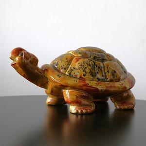 May include: A ceramic turtle ornament with a glossy finish. The turtle has a brown head and legs, and a yellow and brown shell with dark spots. The figurine is posed as if it is walking.