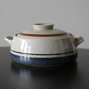 May include: A round ceramic casserole dish with a lid, featuring a speckled off-white glaze, a dark blue band around the base, and a brown ring on the lid. The pot includes two small side handles and a central knob on the lid for easy lifting.