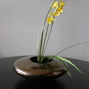 Peut inclure: Un vase rond et bas de couleur bronze avec une ouverture centrale, contenant des orchidées jaunes et du feuillage vert. Le vase est posé sur une surface sombre, créant un contraste avec le fond clair. Arrangement floral moderne.