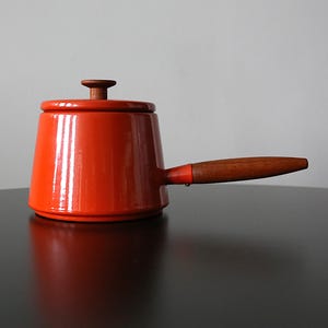 Puede incluir: Una cacerola naranja vibrante con mango y tapa de madera. La olla tiene un diseño clásico, con un cuerpo redondeado y un mango largo y curvo. La tapa presenta un pomo de madera. Este artículo de cocina es perfecto para cocinar.