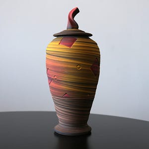 Puede incluir: Urna de cerámica hecha a mano con tapa. La urna presenta un diseño en espiral con bandas horizontales de color, incluyendo tonos de amarillo, naranja y marrón. Se incorporan detalles rojos en forma de diamante. La tapa tiene un asa curvada.