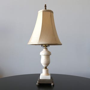 May include: A table lamp featuring a beige fabric bell-shaped shade, a cream-colored urn-shaped base with subtle carved details, and a square metallic base with decorative feet. The lamp is topped with a small, dark brown finial.