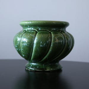 May include: A green ceramic planter with a glossy finish. The planter has a bulbous shape with vertical ridges and a decorative rim. The surface has a crackle effect. It sits on a small, round base. Ideal for plants, it measures approximately 15 cm in diameter.