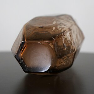 K&ouml;nnte beinhalten: Ein facettiertes, braunes Glasdekorobjekt mit geometrischer Form. Das Objekt hat einen hohlen Innenraum und liegt auf einer dunklen Oberfl&auml;che. Das Glas ist durchscheinend und l&auml;sst Licht durch. Abmessungen nicht sichtbar.