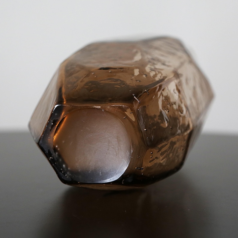 K&ouml;nnte beinhalten: Ein facettiertes, braunes Glasdekorobjekt mit geometrischer Form. Das Objekt hat einen hohlen Innenraum und liegt auf einer dunklen Oberfl&auml;che. Das Glas ist durchscheinend und l&auml;sst Licht durch. Abmessungen nicht sichtbar.