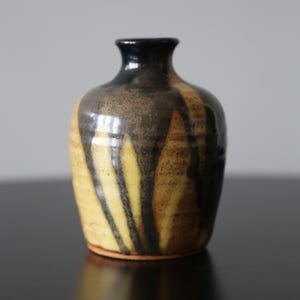 Op de afbeelding: Een kleine, handgemaakte keramische vaas met een afgeronde body en een smalle hals. De vaas heeft een kleurenpalet van geel, bruin en zwart, met donkere druppels die langs de zijkanten naar beneden lopen. De vaas wordt waarschijnlijk voor decoratieve doeleinden gebruikt.