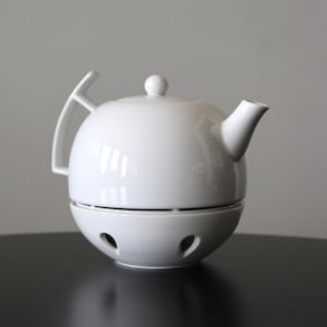 Può includere: Teiera in ceramica bianca con corpo arrotondato, beccuccio piccolo e manico. La teiera poggia su una base abbinata con diversi piccoli fori. Il design è minimalista e moderno, adatto per servire il tè.
