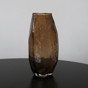K&ouml;nnte beinhalten: Eine braune Glasvase mit strukturierter Oberfl&auml;che und einzigartigem, facettiertem Design. Die Vase hat einen breiten Boden, der sich nach oben verj&uuml;ngt, und steht auf einer dunklen Oberfl&auml;che. Das Glas hat kleine Luftblasen.