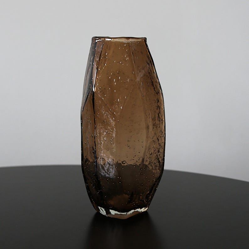 K&ouml;nnte beinhalten: Eine braune Glasvase mit strukturierter Oberfl&auml;che und einzigartigem, facettiertem Design. Die Vase hat einen breiten Boden, der sich nach oben verj&uuml;ngt, und steht auf einer dunklen Oberfl&auml;che. Das Glas hat kleine Luftblasen.
