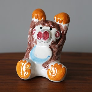 May include: A ceramic figurine of a brown bear with orange paws and a blue and white painted design. The bear is sitting up with its arms raised.
