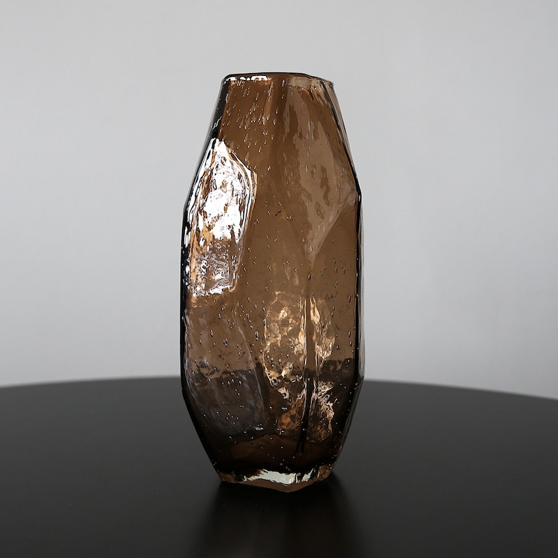 K&ouml;nnte beinhalten: Eine braune Glasvase mit strukturiertem, facettiertem Design. Die Vase hat eine hohe, schlanke Form und steht auf einer schwarzen Oberfl&auml;che. Das Glas hat ein leicht blasenartiges Aussehen.