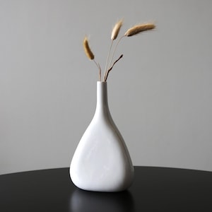 May include: A white ceramic vase with a rounded, triangular shape, holding dried brown grasses.