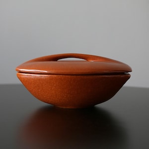 Puede incluir: Un cuenco de cerámica con tapa. El cuenco y la tapa son de color marrón anaranjado moteado. La tapa tiene un asa que se curva sobre la parte superior. El cuenco está sobre una superficie oscura.