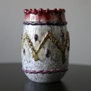 May include: A ceramic vase with a textured, speckled gray body. The top rim is a deep burgundy with a ruffled edge. The vase features gold and purple decorative elements, with a purple wavy band near the base. A unique decorative piece.