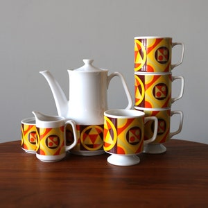 Puede incluir: Un juego de té vintage con una tetera blanca y cuatro tazas. Las tazas y la tetera tienen un patrón geométrico en tonos naranja, amarillo y marrón.