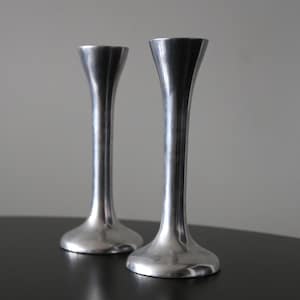 May include: Two silver-colored candle holders with a sleek, modern design. Each holder has a flared top, a slender, elongated body, and a rounded base. The holders are made of a reflective metal and stand on a dark surface.