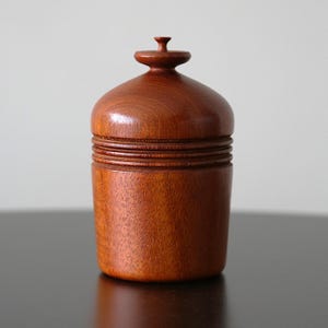 Puede incluir: Un recipiente cilíndrico de madera con un acabado liso de color marrón rojizo. El objeto presenta una tapa redondeada rematada con un pequeño pomo y está decorado con tres líneas horizontales ranuradas que rodean la sección central.