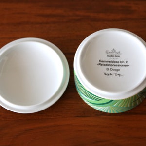 Puede incluir: Una caja de porcelana blanca con un dise&ntilde;o geom&eacute;trico verde y dorado. La tapa de la caja tiene el texto "Sammeldose Nr. 2 "Reiseimpressionen" B. Doege" escrito en ella.