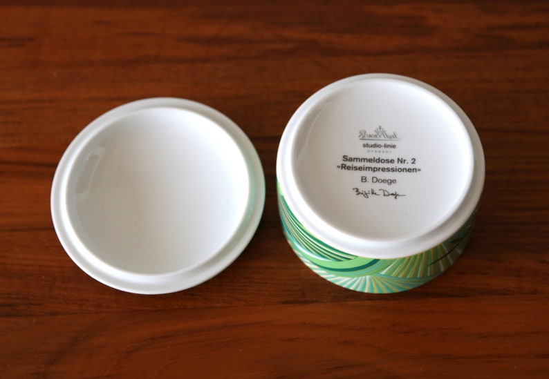 Puede incluir: Una caja de porcelana blanca con un dise&ntilde;o geom&eacute;trico verde y dorado. La tapa de la caja tiene el texto "Sammeldose Nr. 2 "Reiseimpressionen" B. Doege" escrito en ella.