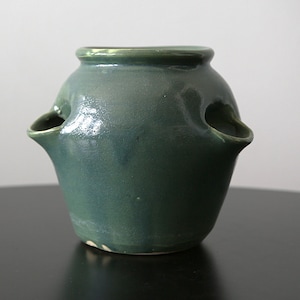 May include: A handcrafted, teal-colored ceramic vase with two spout-like openings on the sides. The vase has a rounded body and a slightly flared rim. The glaze has a glossy finish, and the vase sits on a dark surface.