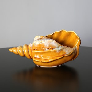 May include: A ceramic bowl shaped like a conch shell with a glossy finish. The exterior features a vibrant mustard yellow glaze with subtle white and light brown highlights along the ridges and the spiral tip, creating a smooth, reflective surface.